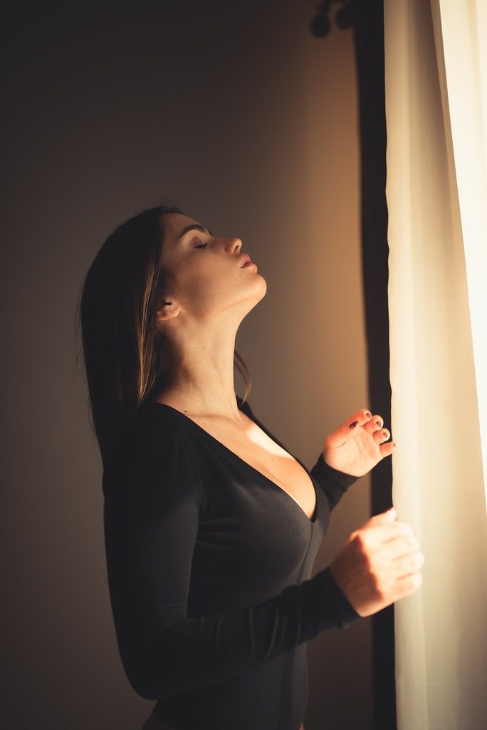 A woman pauses by a sunlit window, eyes closed, taking a calm moment for herself.